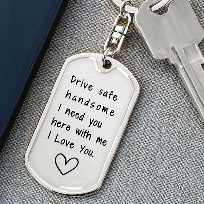 Surgical steel military dog tag keychain with swivel top, engraved with 'Drive Safe Handsome, I Need You Here with Me, I Love You,' and a heart outline, placed diagonally on a gray mat.